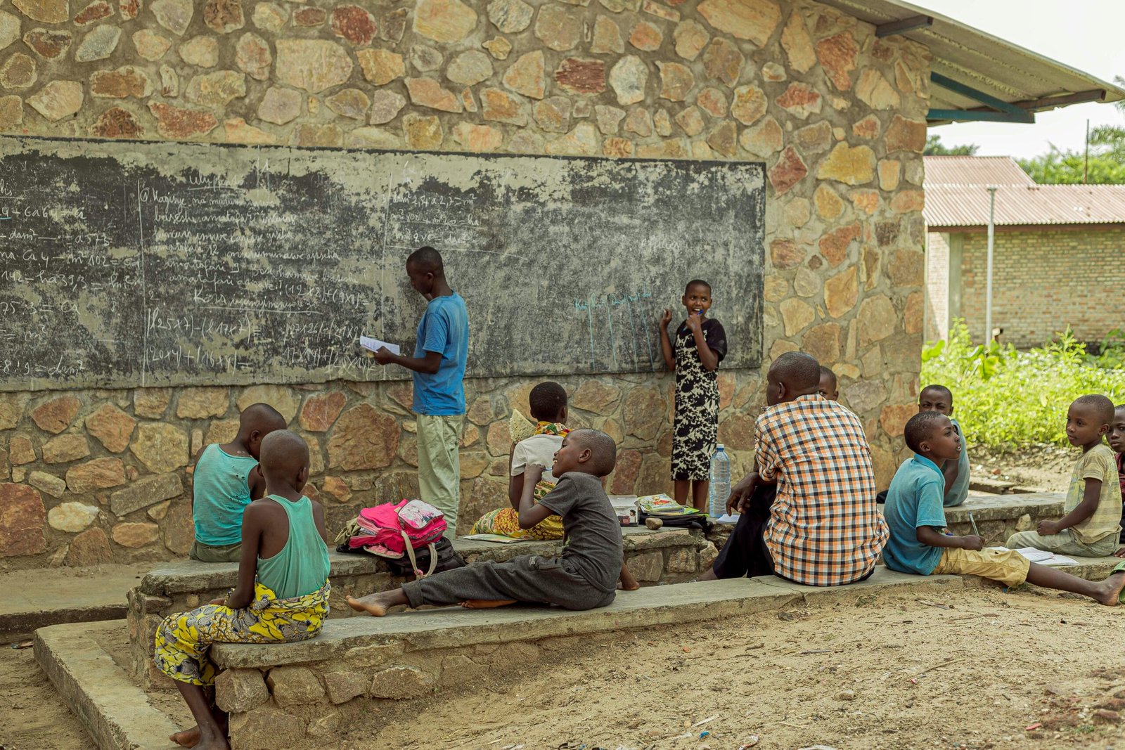 Children studying outdoors with a teacher, engaging with a blackboard at a rural school.